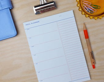 Pode incluir: Um planeador de refeições branco com linhas azuis e rosa. O planeador tem um clipe na parte superior e as palavras "MEAL PLANNER" estão impressas na parte superior. O planeador está sobre uma superfície de madeira com um lápis e um pequeno prato de clipes de papel coloridos.