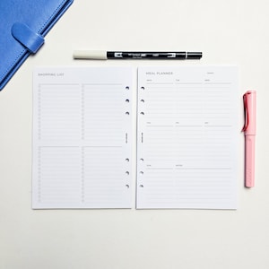 May include: A white planner with a shopping list and meal planner section. The planner has a hole punch design for a binder. The planner is on a white surface with a black and white marker and a pink pen.