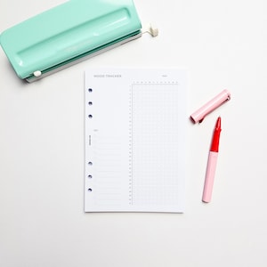 a mood tracking notebook, a green stapler, and a red pen on a white background.