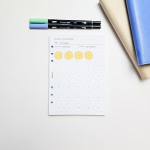 May include: A white 30-day challenge planner with the goal "No Spend" and start and finish dates. The planner has a grid of circles, some filled in yellow. Two black pens with green and blue tips are on top, along with a beige and blue notebook.