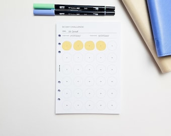 May include: A white 30-day challenge planner with the goal "No Spend" and start and finish dates. The planner has a grid of circles, some filled in yellow. Two black pens with green and blue tips are on top, along with a beige and blue notebook.
