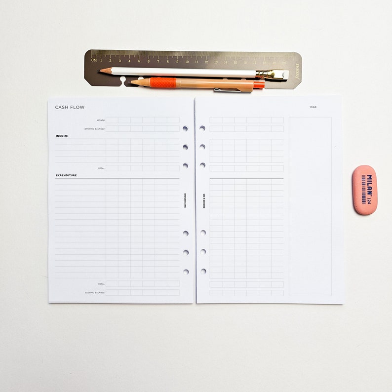 May include: A white cash flow planner with a black grid and the words "CASH FLOW" at the top. The planner is open to a page with the headings "INCOME" and "EXPENDITURE".