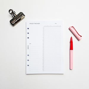 a mood tracking notebook, a red pen, and a black binder clip on a white background.