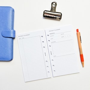 May include: Two white project planner pages with a black header that says "Project Planner". The left page has a column for "Date" and "Task". The right page has sections for "Description", "Start Date", "End Date", "Priority", "Budget", "Milestones", and "Notes".