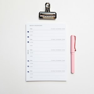 May include: A white book inventory tracker with a silver binder clip and a pink pen. The tracker has spaces to write the title, author, and genre of books. It also has checkboxes for "to read", "in progress", and "done".
