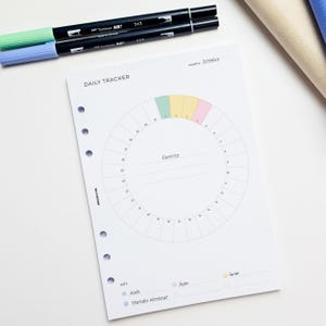 May include: A white daily tracker planner page with a circular exercise chart, color-coded for activities like walking and gym. Includes the text "DAILY TRACKER" and "October". Two black Tombow ART markers are on top.