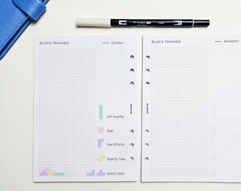 May include: Two open planner pages with grid lines, labeled "Block Tracker" for October and November. A black and white pen rests above the pages. The left page has color-coded blocks for tracking habits like "Eat Healthy" and "Bubble Bath."