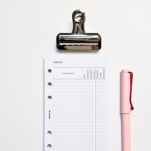 May include: A top-down shot of a silver binder clip, a white planner page with the word "INBOX" and columns, and a pink pen with a red cap. The planner page has columns labelled "Next", "Schedule", "Projects", "Waiting", "Someday", "Maybe", and "Reference".