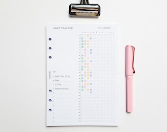 May include: A white habit tracker with a month of October calendar, colorful dots, and a key for tracking habits. A pink pen rests beside the tracker, and a silver binder clip is at the top. The tracker has a list of habits to track.
