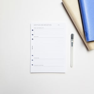 May include: A white gratitude and reflection journal with a black pen on a white surface. The journal has a section for listing things you are grateful for, a reflection section, a section for affirmations, and a section for tomorrow's intentions.