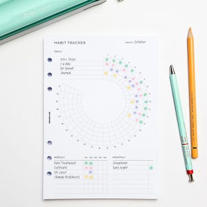 May include: A white habit tracker planner page with a circular design for daily habits and sections for weekly and monthly goals. Includes text "HABIT TRACKER" and "October". Two pens, one orange and one teal, are next to the page.