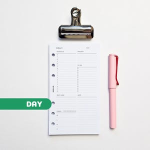 May include: A white planner page with a daily schedule, priority list, and to-do section. A pink pen with a red clip and a silver binder clip are next to the page. The word "DAY" is on a green bar.
