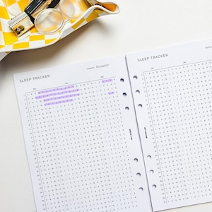 May include: Two open pages of a sleep tracker planner with the text "SLEEP TRACKER" at the top. The December section is highlighted in purple. A yellow and white checkered tray with office supplies is in the background.