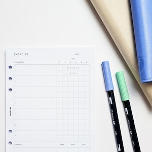 May include: A white exercise log with a grid for tracking strength training workouts. The grid has columns for set, weight, and reps. The log also has sections for cardio, time, distance, and calories burned. Two black markers with light blue and light green caps are on the right side of the page.