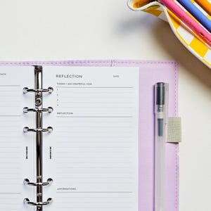 May include: A lavender planner with a silver binder mechanism, open to a page titled "REFLECTION." The page includes prompts for gratitude and affirmations. A clear pen with a black cap is attached to the planner. A yellow and white dish with colored pencils is in the background.