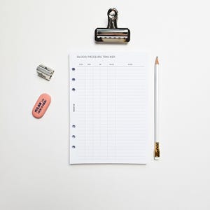 May include: A white blood pressure tracker sheet with a lined grid, a pencil, a pink eraser, a silver pencil sharpener, and a silver binder clip. The sheet has the words "BLOOD PRESSURE TRACKER" printed at the top.