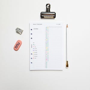 May include: A white daily tracker planner with a grid for tracking spending, a pencil, a pink eraser, a silver binder clip, and a pencil sharpener. The planner has a key with coloured dots and the text "DAILY TRACKER".