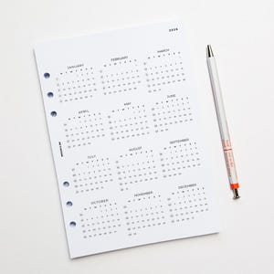 May include: A white paper calendar page for the year 2026 featuring twelve monthly grids with black text and numbers. The page has five circular punch holes along the left edge and the text Badger & Ink. A white pen with orange accents lies beside it.
