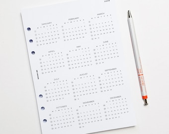 May include: A white paper calendar page for the year 2026 featuring twelve monthly grids with black text and numbers. The page has five circular punch holes along the left edge and the text Badger & Ink. A white pen with orange accents lies beside it.