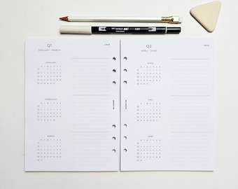 May include: A white planner with black text and a calendar for January through June. The planner is divided into two sections, one for Q1 and one for Q2. The planner has a hole punch on the left side and a spiral binding on the right side.