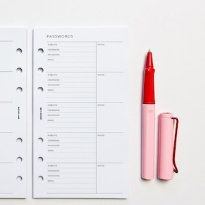 May include: A white, 6-ring binder with a black header that reads "PASSWORDS". The binder has five sections for website, username, password, email, and notes. A red and pink pen is lying on top of the binder.
