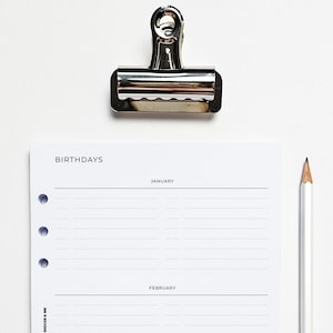 May include: A white page with the word "BIRTHDAYS" at the top, followed by the months "JANUARY" and "FEBRUARY". The page is divided into two sections, one for each month. There are lines for writing down birthdays. A silver binder clip is holding the page to a white surface. A white pencil is lying on the surface.