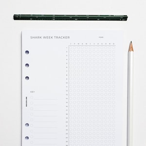 May include: A white Shark Week Tracker planner page with a grid for tracking, a pencil, and a ruler. The planner page has the text "SHARK WEEK TRACKER" at the top and a key on the left side. The ruler is dark green.