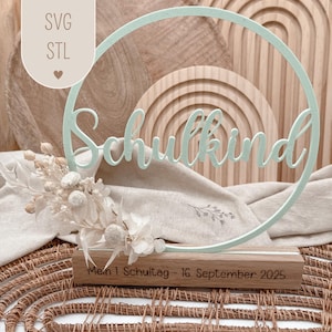 Könnte beinhalten: Minzgrünes Holzschild mit dem Wort "Schulkind" in Schreibschrift, in einem kreisförmigen Rahmen. Das Schild steht auf einem Holzsockel mit dem Text "Mein 1 Schultag - 16. September 2025". Trockenblumen schmücken die Basis.