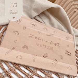 Puede incluir: Una tabla de cortar de madera de color claro con ilustraciones grabadas de un lápiz, nubes, arcoíris y otras formas. El texto "Ich rechne mir die Welt, WIE SIE MIR GEFÄLLT" está grabado en la tabla. La tabla es rectangular.