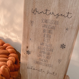 Puede incluir: Un letrero de madera con la palabra "Winterzeit" en escritura cursiva. Debajo, una lista de palabras y frases con temática invernal, como "copos de nieve" y "calcetines acogedores". Copos de nieve y corazones decorativos.