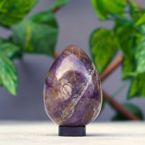 May include: A purple amethyst crystal egg with white and brown swirls, sitting on a black stand.