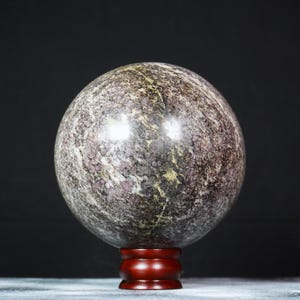 May include: A polished, spherical stone orb featuring a mottled pattern of deep purple, dark gray, and earthy brown tones with thin, light-colored mineral veins throughout. The sphere rests securely on a small, circular, dark reddish-brown wooden stand.