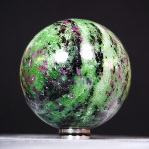 Può includere: Una pietra sferica lucida caratterizzata da un mix vibrante di zoisite verde brillante, inclusioni di rubino rosa magenta intenso e striature di orneblenda nera. La superficie è liscia e riflettente, con un motivo minerale naturale.