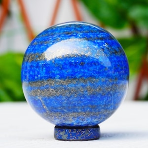 Puede incluir: Una esfera azul pulida con motas doradas, sobre una pequeña base a juego. La bola de piedra preciosa tiene un color azul intenso con bandas doradas y grises. La esfera es probablemente de lapislázuli.
