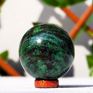 Könnte beinhalten: Eine grüne und schwarze Edelsteinkugel auf einem roten Ständer. Die Kugel ist poliert und hat eine glatte Oberfläche.
