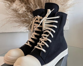 Könnte beinhalten: Schwarze High-Top-Canvas-Sneaker mit weißen Schnürsenkeln und einer weißen Gummisohle. Die Schuhe stehen auf einem grauen Karton.