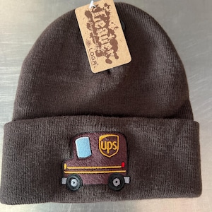 May include: Brown knit beanie with a brown UPS truck embroidered on the front. The truck has a yellow "UPS" logo and a blue windshield.