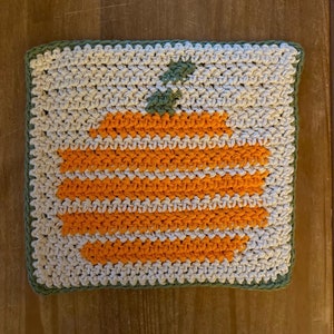 May include: A crocheted dishcloth with a pumpkin design in orange and white on a beige background. The dishcloth has a green border.