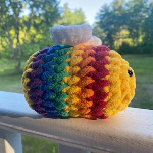 May include: A plush, crocheted bee toy featuring a vibrant rainbow-striped body with bands of purple, blue, green, yellow, orange, and red yarn. The bee has two small, rounded white wings on its back and a single black bead eye visible on the side.