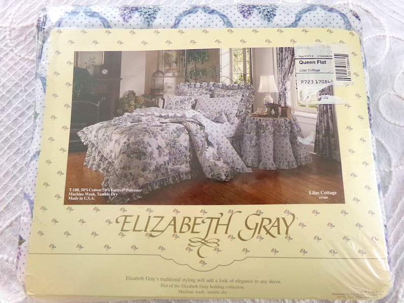 May include: A white and purple floral queen-sized flat sheet with a ruffled edge. The sheet is part of the Elizabeth Gray Lilac Cottage bedding collection. The packaging includes the product name, size, and care instructions.