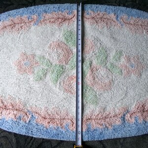 May include: A white oval bath mat with a blue and pink floral design. The mat has a fluffy texture and is approximately 24 inches long.