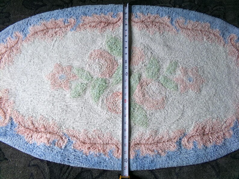 May include: A white oval bath mat with a blue and pink floral design. The mat has a fluffy texture and is approximately 24 inches long.