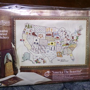 May include: A framed Paragon Needle Craft kit featuring a map of the United States, embroidered with state symbols. The kit includes the text "America The Beautiful" and "One Nation Under God". The packaging includes the words "creative crewel stitchery".