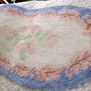 May include: A white oval bath mat with a blue border and pink floral design. The mat has a soft, fluffy texture.