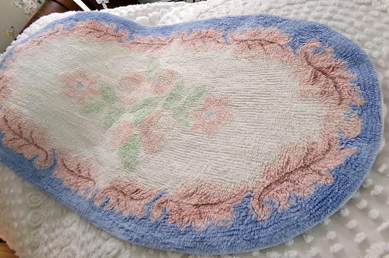 May include: A white oval bath mat with a blue border and pink floral design. The mat has a soft, fluffy texture.