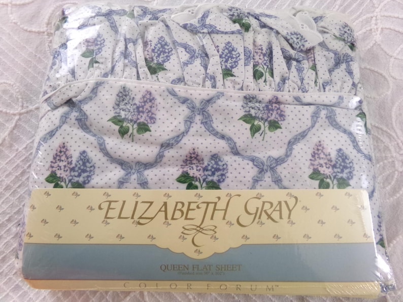May include: A white queen flat sheet with a lilac floral pattern and blue ribbon design. The sheet is packaged and labeled "Elizabeth Gray" and "Colorforum".