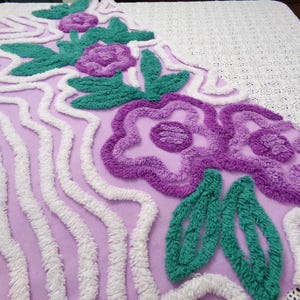 May include: A lilac-colored, tufted rug with a floral design. The rug features purple flowers, green leaves, and white wavy lines. The rug is likely made of a soft, plush material, and is designed for decorative purposes.