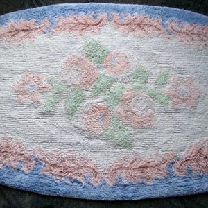 May include: Oval shaped bath mat with a white background and a floral design in pink, green, and blue. The border is a light blue with a pink trim.