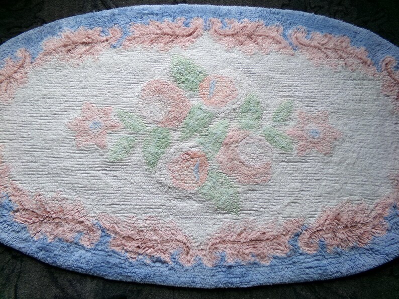 May include: Oval shaped bath mat with a white background and a floral design in pink, green, and blue. The border is a light blue with a pink trim.