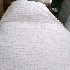 May include: A white bedspread featuring a raised tufted texture in a repeating circular and geometric pattern. The fabric is solid white with a consistent, dense arrangement of small, soft, rounded tufts that create a tactile, textured surface.